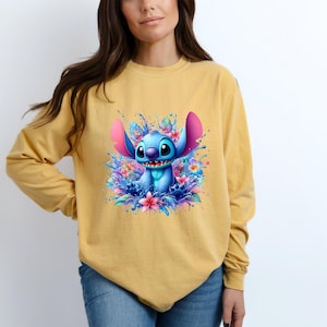 May include: Mustard yellow long-sleeve shirt featuring a blue cartoon character with large ears and a wide smile, surrounded by colorful flowers and water splashes. The character is the focal point of the graphic design.