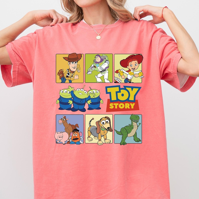 May include: A coral-colored t-shirt featuring a colorful graphic of characters from the animated film "Toy Story." The design includes Woody, Buzz Lightyear, Jessie, the aliens, Mr. Potato Head, Slinky Dog, and Rex, with the "Toy Story" logo.