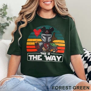 May include: Forest green t-shirt featuring a graphic of a character in Mandalorian armor with Mickey Mouse ears, holding a red heart balloon, and Grogu. The design is set against a sunset background with the text "THIS IS THE WAY".