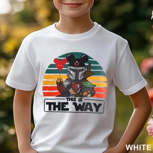May include: White t-shirt featuring a cartoon graphic of The Mandalorian with a Mickey Mouse helmet and Grogu. The design includes a red heart-shaped balloon and a sunset background with the text "THIS IS THE WAY".