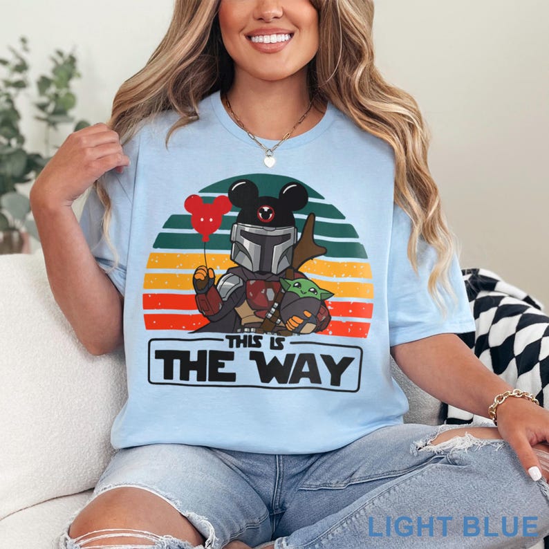 May include: Light blue t-shirt featuring a graphic of a Mandalorian character with Mickey Mouse ears, holding a red heart balloon, and Grogu. The design is set against a sunset backdrop with the text "THIS IS THE WAY".