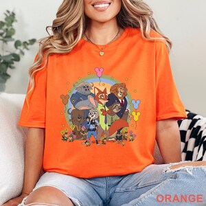 Puede incluir: Camiseta naranja brillante con un dise&ntilde;o de dibujos animados de personajes de animales, un arco&iacute;ris y flores. La palabra "ORANGE" est&aacute; en la parte inferior. La camiseta es de color liso y parece estar hecha de un material suave.