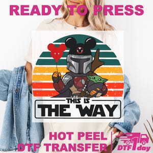 May include: White t-shirt featuring a cartoon design of The Mandalorian with a Mickey Mouse helmet and Grogu. The design includes a red balloon, a sunset background, and the text "THIS IS THE WAY". The words "READY TO PRESS" and "HOT PEEL DTF TRANSFER" are also visible.