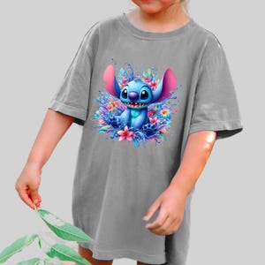 May include: Gray t-shirt featuring a vibrant illustration of Stitch from Lilo & Stitch. The blue character is surrounded by colorful flowers and water splashes. The shirt is a comfortable, oversized fit.