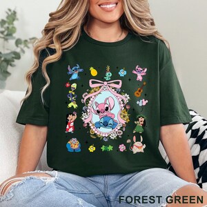 May include: Forest green t-shirt with a colourful graphic of Stitch and Angel characters. The design includes a central oval with the two characters, surrounded by flowers and other themed elements. The text "FOREST GREEN" is printed at the bottom.