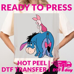 May include: A light-coloured t-shirt featuring a cartoon image of Eeyore and Piglet. Eeyore is blue with a pink bow, and Piglet is pink. The text "READY TO PRESS", "HOT PEEL", and "DTF TRANSFER" are displayed in pink.