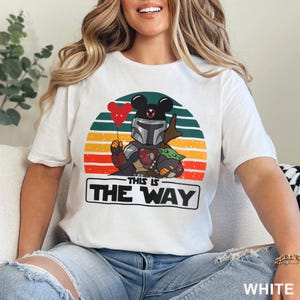 May include: White t-shirt featuring a graphic of a Mandalorian character with Mickey Mouse ears, holding a red heart balloon, and Grogu. The design is set against a sunset background with the text "THIS IS THE WAY" in a rectangular box.