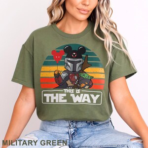 May include: Military green t-shirt featuring a graphic of The Mandalorian with Mickey Mouse ears, holding Grogu and a red heart balloon. The text "THIS IS THE WAY" is below the graphic, set against a sunset background.