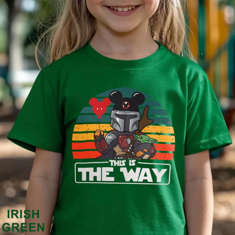 May include: Irish green t-shirt featuring a graphic of The Mandalorian with a Mickey Mouse-themed helmet and Baby Yoda. The design includes a red balloon and the text "THIS IS THE WAY" in a white rectangle. The shirt is for children.