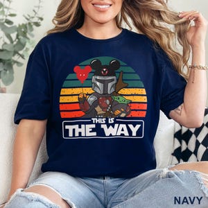 May include: Navy blue t-shirt featuring a graphic of a Mandalorian character with Mickey Mouse ears, holding a red heart balloon, and Grogu. The design is set against a sunset backdrop with the text "THIS IS THE WAY".