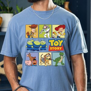May include: Light blue t-shirt featuring a colorful graphic of Toy Story characters in square panels. The design includes Woody, Buzz Lightyear, Jessie, the Aliens, and others, with the words "TOY STORY" in red and yellow.
