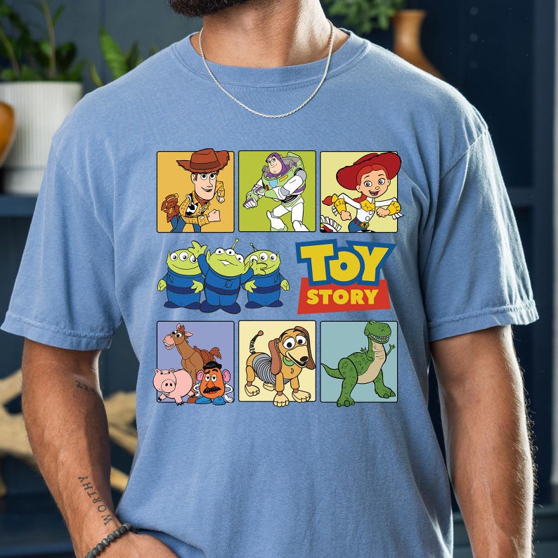 May include: Light blue t-shirt featuring a colorful graphic of Toy Story characters in square panels. The design includes Woody, Buzz Lightyear, Jessie, the Aliens, and others, with the words "TOY STORY" in red and yellow.