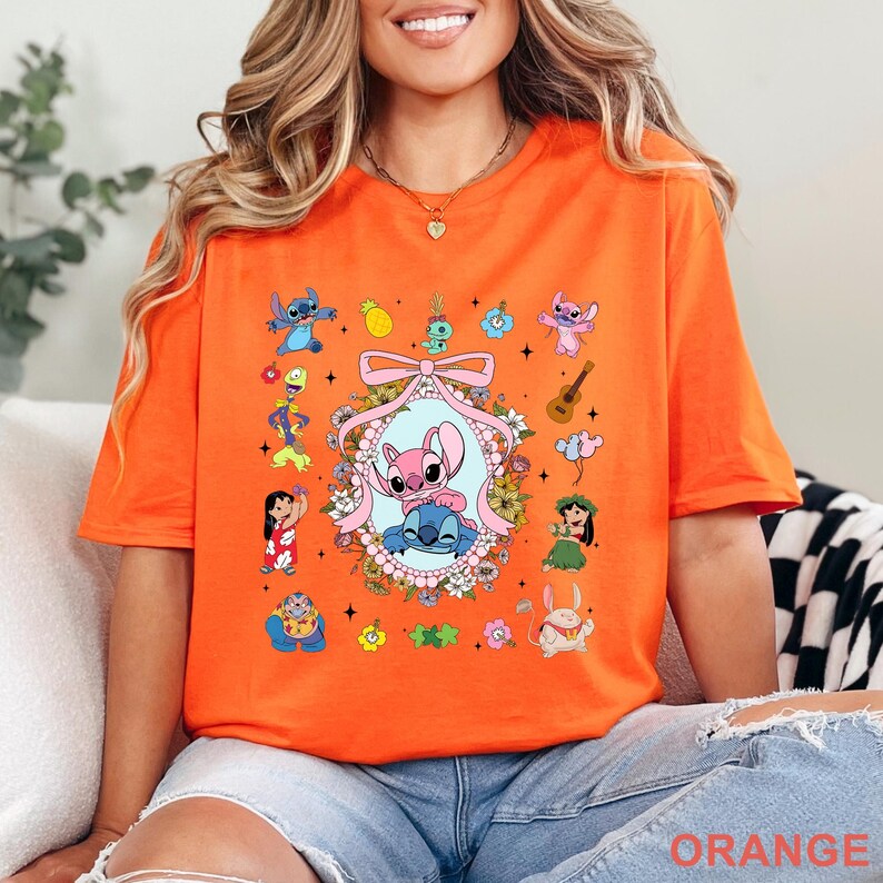 May include: Bright orange t-shirt with a vibrant graphic of Stitch and Angel, set within a floral and tropical arrangement. The design includes a central oval frame with the two characters. The word "ORANGE" is printed at the bottom.