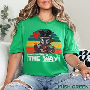May include: Irish green t-shirt featuring a graphic of a Mandalorian-inspired character with Mickey Mouse ears, holding a red heart balloon, and Grogu. The design is set against a sunset backdrop with the text "THIS IS THE WAY".
