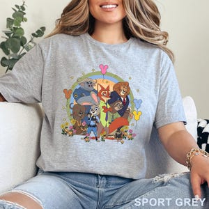 Puede incluir: Camiseta gris deportivo con un gr&aacute;fico colorido de personajes de Zootopia. El dise&ntilde;o incluye a Judy Hopps, Nick Wilde y otros personajes en un arreglo circular con flores, globos y un arco&iacute;ris. La camiseta est&aacute; hecha de un material suave.