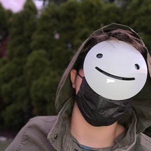 May include: A person wearing a green hooded jacket and a black face mask. The person is wearing a white mask with a smiling face.