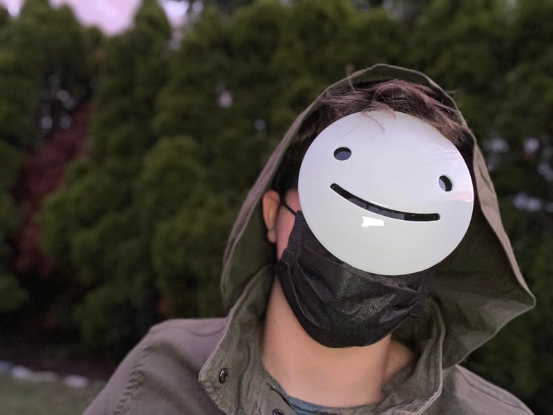 May include: A person wearing a green hooded jacket and a black face mask. The person is wearing a white mask with a smiling face.