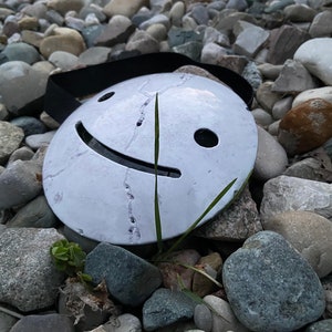 May include: A white, round mask with a smiling face and a black strap. The mask is lying on a bed of gray rocks.