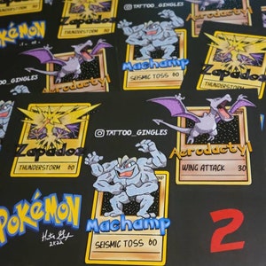 May include: A collection of Pokemon trading cards featuring illustrations of Zapdos, Aerodactyl, and Machamp. The cards are black with yellow borders and feature the Pokemon's name and attack power. The cards are signed "Hita Sif 2K22".