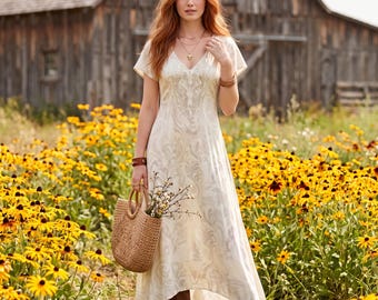 May include: A cream-colored midi dress with a subtle swirling pattern, short sleeves, and a V-neckline. The dress features delicate embroidery around the neckline and is paired with brown leather boots and a woven straw handbag with round handles.