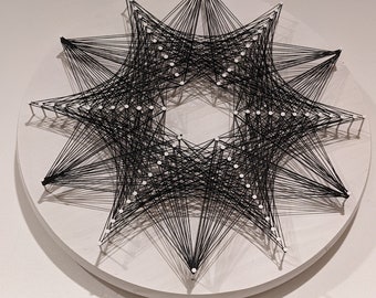 May include: A black string art piece in a starburst design. The artwork features black thread stretched between white pins on a white circular wooden base. The geometric pattern creates a striking visual effect, suitable for wall decor.