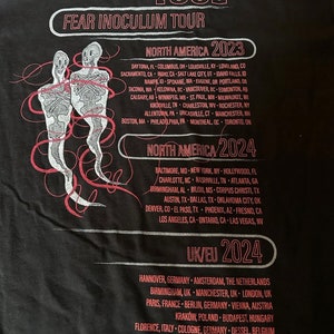 May include: Black t-shirt with white text listing the dates and locations for the Tool Fear Inoculum Tour in North America and Europe in 2023 and 2024.
