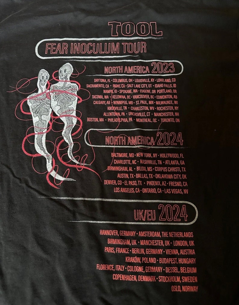 May include: Black t-shirt with white text listing the dates and locations for the Tool Fear Inoculum Tour in North America and Europe in 2023 and 2024.