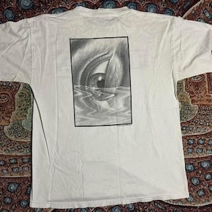 Puede incluir: Camiseta blanca con un estampado gráfico en blanco y negro de un ojo mirando hacia arriba desde el agua. El gráfico está en un marco rectangular.