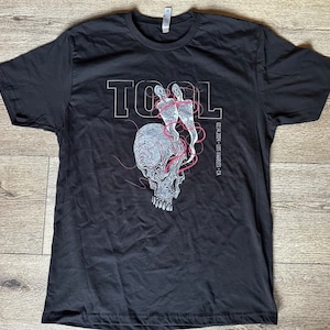 May include: Black t-shirt featuring a white graphic of a skull with red lines and the word "TOOL" in white lettering. The text "02.24.2014 - Los Angeles, CA" is printed in white below the graphic.