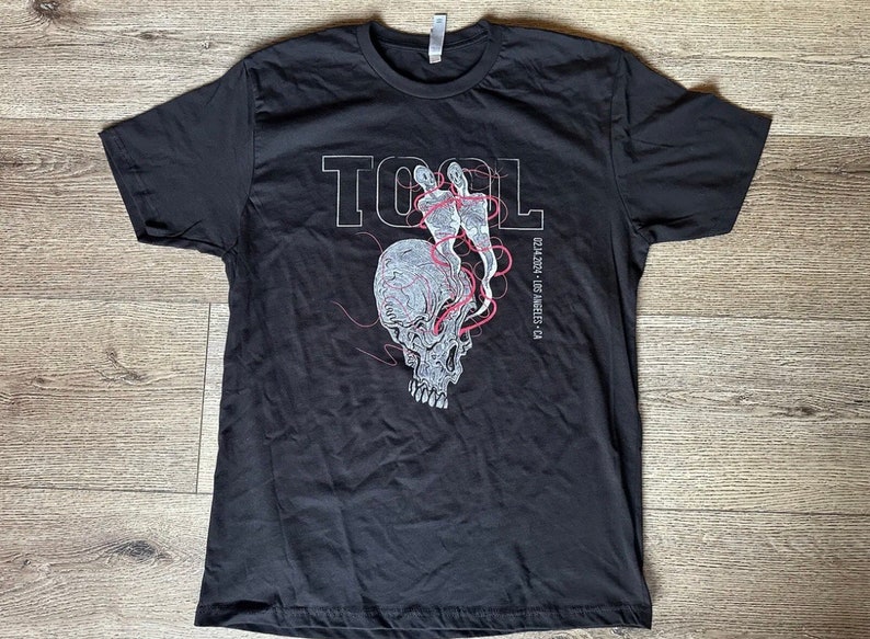 May include: Black t-shirt featuring a white graphic of a skull with red lines and the word "TOOL" in white lettering. The text "02.24.2014 - Los Angeles, CA" is printed in white below the graphic.