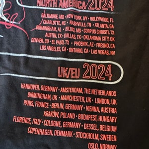 May include: A black t-shirt with red text listing tour dates for North America and Europe in 2024. The text includes cities in the United States, Canada, Germany, the Netherlands, United Kingdom, France, Austria, Poland, Hungary, Italy, Belgium, Denmark, Sweden, and Norway.