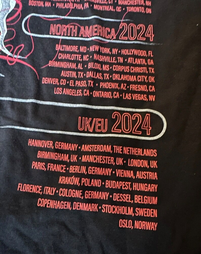 May include: A black t-shirt with red text listing tour dates for North America and Europe in 2024. The text includes cities in the United States, Canada, Germany, the Netherlands, United Kingdom, France, Austria, Poland, Hungary, Italy, Belgium, Denmark, Sweden, and Norway.
