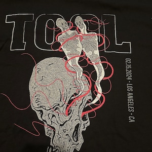 May include: Black t-shirt with a white graphic of a skull with two skeletons inside. The skeletons are connected by red lines. The word "TOOL" is printed in white above the skull. The text "02.15.2024 - LOS ANGELES - CA" is printed in white on the right side of the shirt.