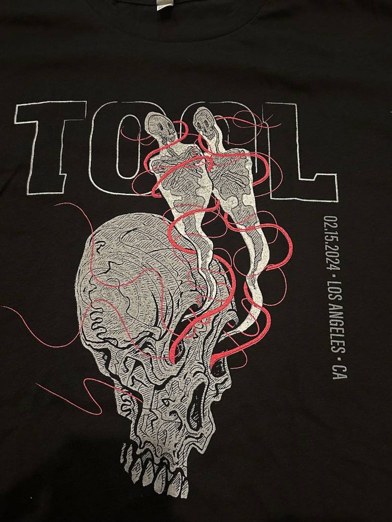 May include: Black t-shirt with a white graphic of a skull with two skeletons inside. The skeletons are connected by red lines. The word "TOOL" is printed in white above the skull. The text "02.15.2024 - LOS ANGELES - CA" is printed in white on the right side of the shirt.