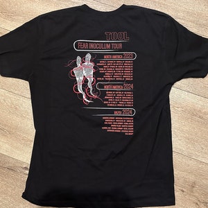 May include: Black t-shirt with a white graphic of two figures and the text "TOOL FEAR INOCULUM TOUR" with tour dates for North America in 2023 and 2024 and the United Kingdom in 2024.