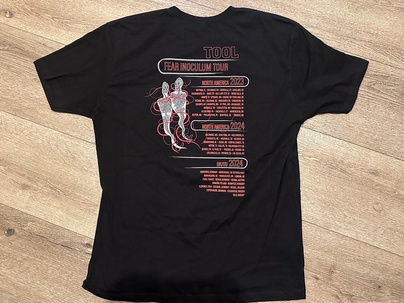 May include: Black t-shirt with a white graphic of two figures and the text "TOOL FEAR INOCULUM TOUR" with tour dates for North America in 2023 and 2024 and the United Kingdom in 2024.