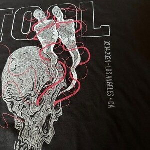 May include: Black t-shirt featuring a white skull graphic and two skeletons with red lines. The text "TOOL" is printed in white on the top left and "02.14.2024 - LOS ANGELES - CA" is printed in white on the right side.