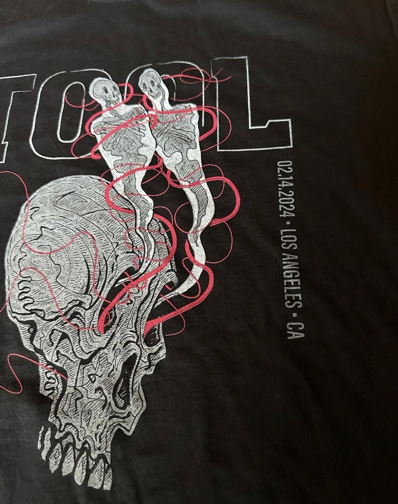 May include: Black t-shirt featuring a white skull graphic and two skeletons with red lines. The text "TOOL" is printed in white on the top left and "02.14.2024 - LOS ANGELES - CA" is printed in white on the right side.