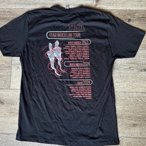 May include: Black t-shirt with a white graphic of two figures and the text "TOOL FEAR INOCULUM TOUR" with tour dates for North America and the UK in 2023 and 2024.