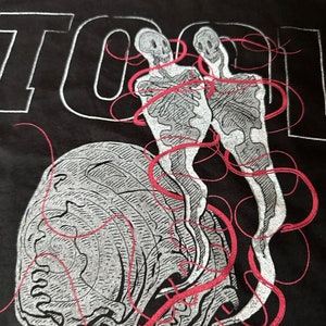 May include: Black t-shirt featuring a white and red graphic design of two skeletons with red lines connecting them. The word "TOOL" is printed in white at the top of the shirt.
