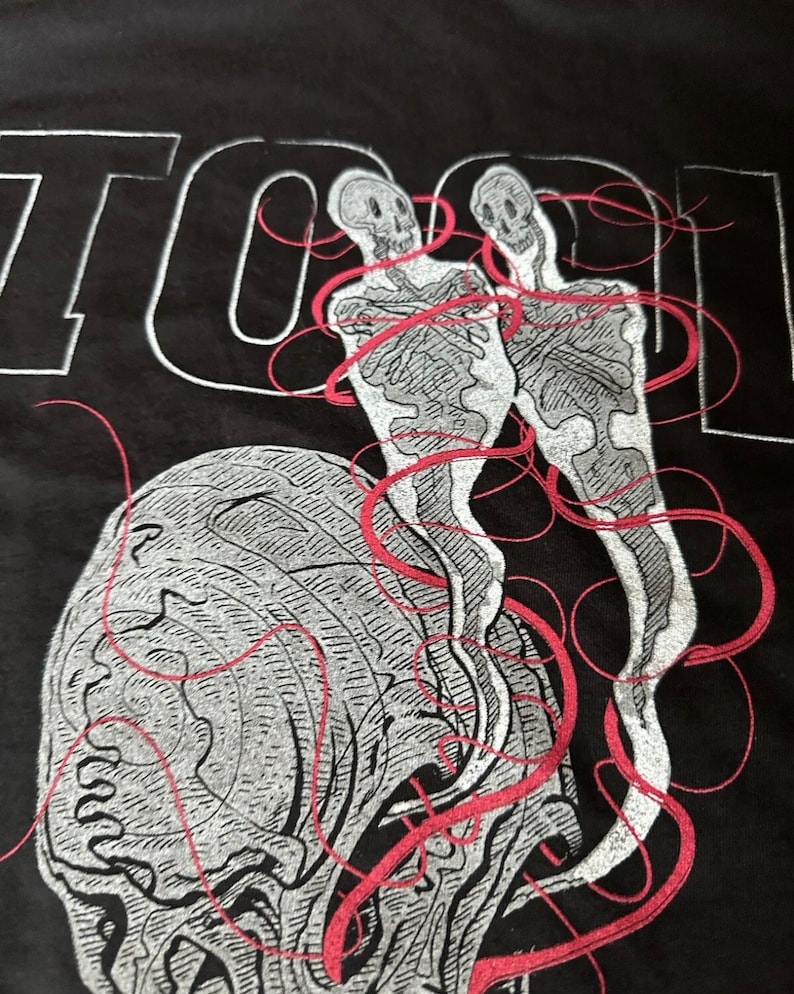 May include: Black t-shirt featuring a white and red graphic design of two skeletons with red lines connecting them. The word "TOOL" is printed in white at the top of the shirt.