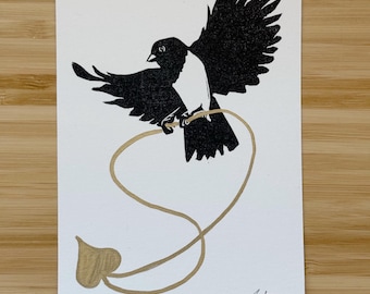 Op de afbeelding: Een zwart-wit vogelillustratie met uitgespreide vleugels, zittend op een goudkleurige hartvormige lijn. Het woord "Love" staat eronder, met de handtekening van de kunstenaar en het jaar '25. De illustratie staat op een witte rechthoekige kaart.