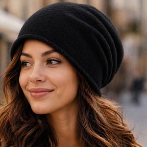 May include: A black beanie hat with the Alster Herz logo. The hat is worn by a person wearing a tan trench coat and a white shirt. The background is a blurred outdoor scene.