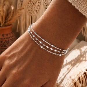 Op de afbeelding: Een zilveren armbandenset met drie fijne strengen. De bovenste streng is een fijne ketting, de middelste heeft kleine, langwerpige kralen en de onderste kleine ronde kralen. De armbanden worden om een pols gedragen.