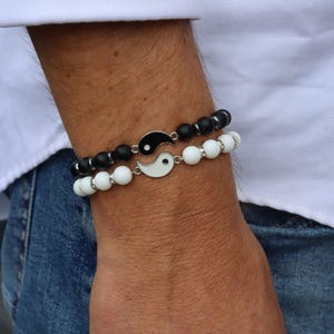 Puede incluir: Dos pulseras de cuentas a juego con un símbolo del Yin Yang. Una pulsera tiene cuentas negras, la otra blancas. El símbolo del Yin Yang es blanco y negro con un borde plateado. Las pulseras se llevan en la muñeca.