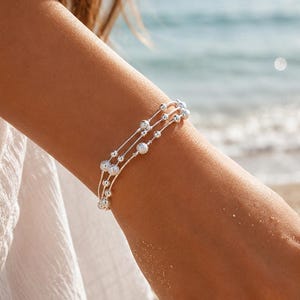 Può includere: Un bracciale d'argento con tre fili, ognuno ornato da piccole perle rotonde di diverse dimensioni. Il bracciale è indossato su un polso, con una spiaggia e il mare sullo sfondo. Un gioiello delicato.
