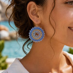 Op de afbeelding: Close-up van een vrouw die grote, ronde oorbellen draagt. De oorbellen hebben een cirkelvormig ontwerp met concentrische ringen van blauwe kralen en gouden accenten. De oorbellen zijn opgehangen aan goudkleurige haken.