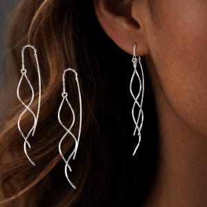 Könnte beinhalten: Lange, silberfarbene Ohrringe mit einem zarten, wellenförmigen Design. Die Ohrringe haben eine Kette, die durch das Ohr geht, und ein fließendes, verdrehtes Metallelement. Die Ohrringe werden an einer Person gezeigt.