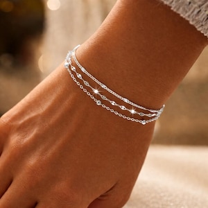 Puede incluir: Un brazalete plateado en una muñeca. El brazalete presenta tres delicadas cadenas: una cadena de cuentas, una cadena con pequeños eslabones ovalados y una cadena con un diseño texturizado. El brazalete es una joya.