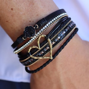 Könnte beinhalten: Mehrere schwarze Armbänder mit goldenen und silbernen Akzenten. Ein Armband hat einen goldenen Herzanhänger. Andere Armbänder haben Strass- und Kristallverzierungen. Die Armbänder sind geschichtet und am Handgelenk getragen.